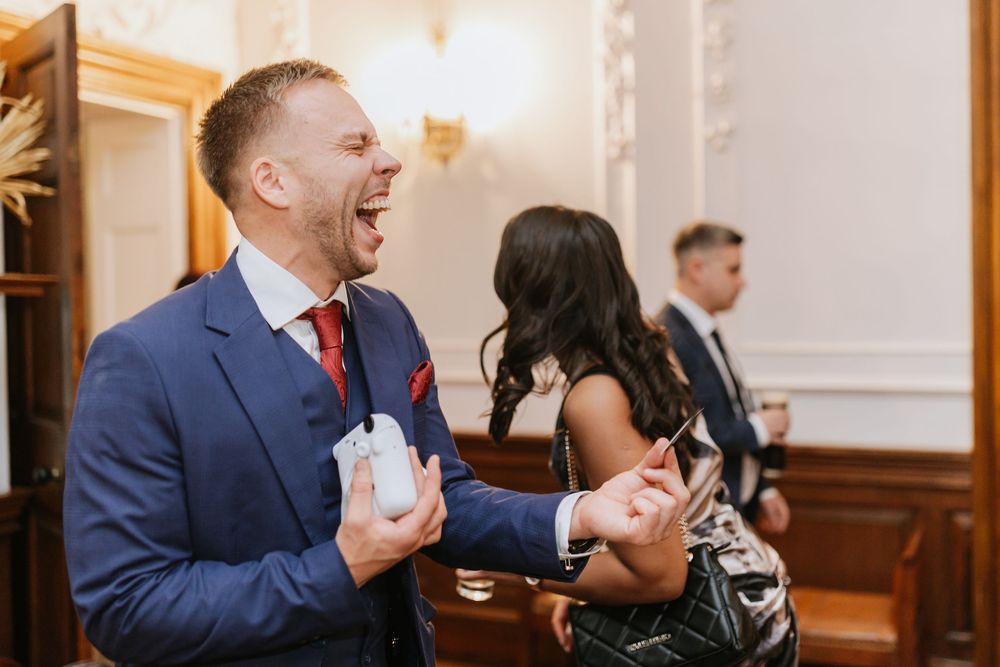 A person in a navy suit laughs joyfully while holding a drink at an indoor event.