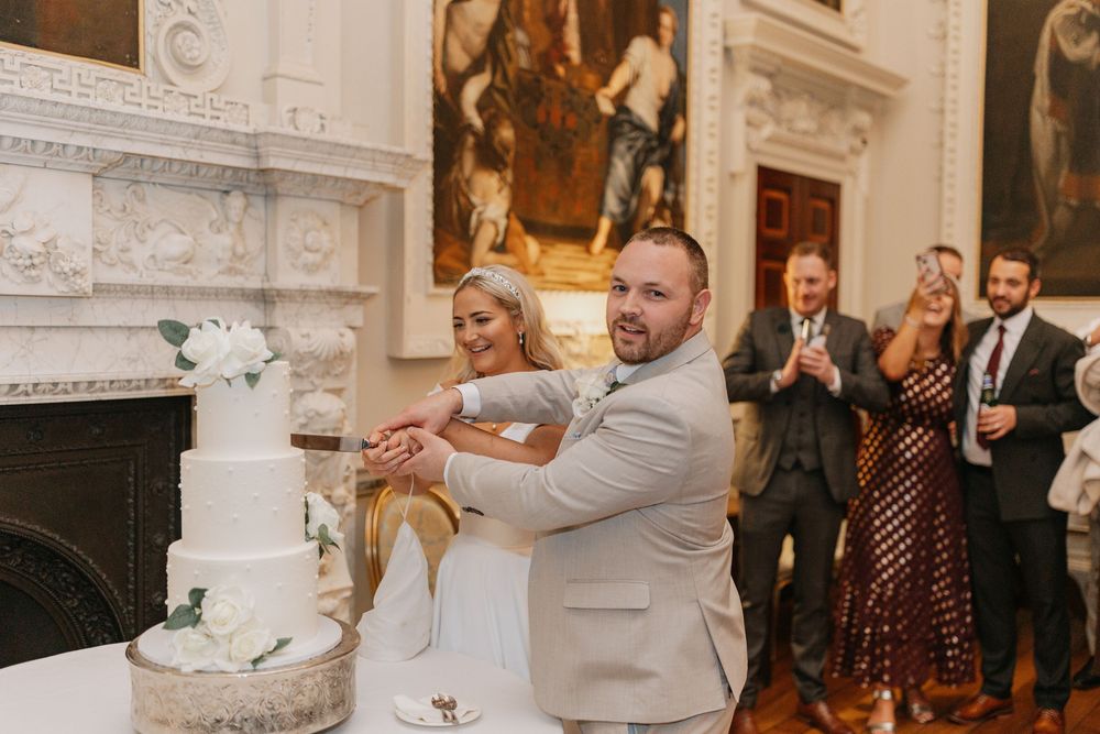 Wedding couple cuts their elegant white tiered cake at an ornate venue during their reception celebration.