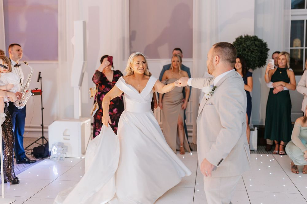 A couple shares their first dance at a wedding reception in an elegant white room with guests watching.
