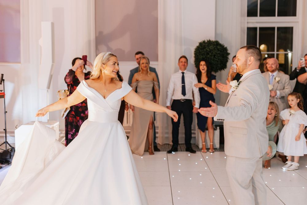 Wedding guests dancing and celebrating in a bright white ballroom with large windows.