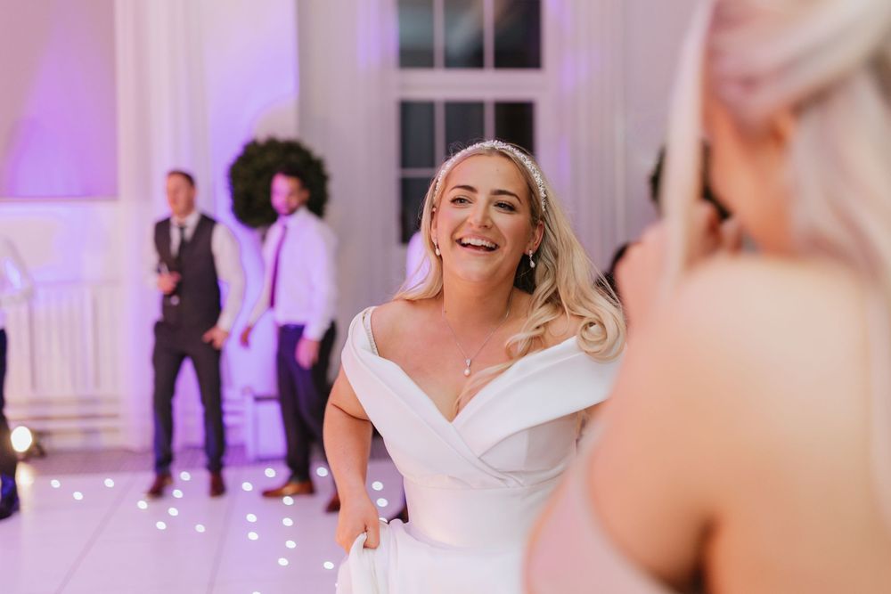 A bride in a white off-shoulder wedding dress smiles joyfully on the dance floor during her wedding reception.