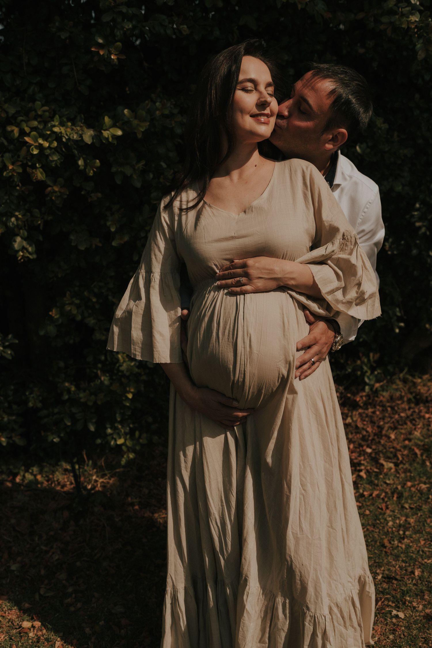 A romantic maternity photoshoot in nature with a couple wearing light colored clothing embracing in warm sunlight.