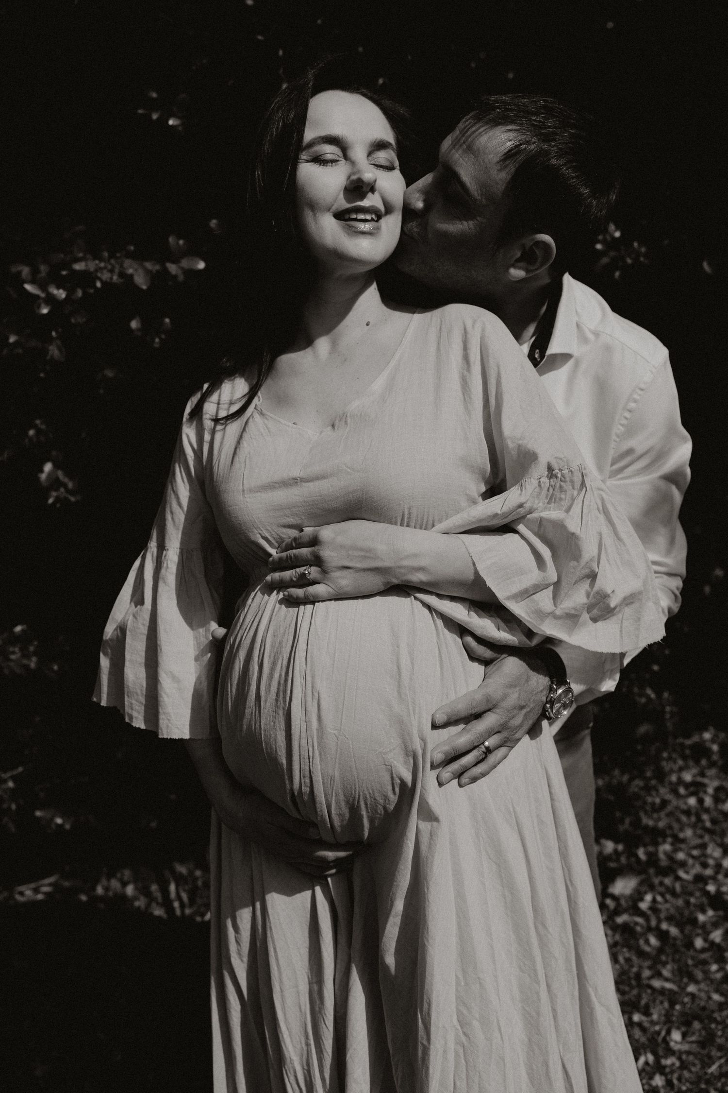 Black and white maternity photos showing tender moments between an expectant couple in flowing white attire.