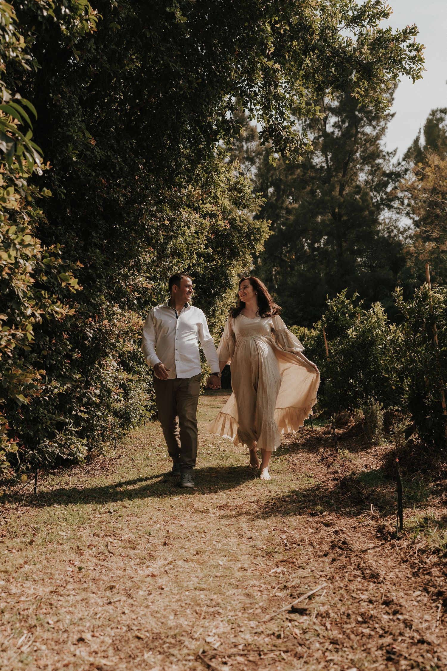 Romantic outdoor photoshoot of a couple strolling along a tree-lined path in neutral toned clothing.