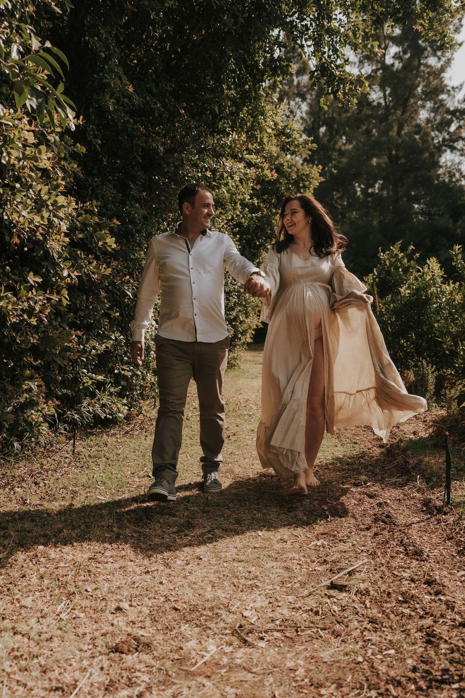 Couple walking hand in hand through a sunlit path wearing coordinated cream and white outfits.