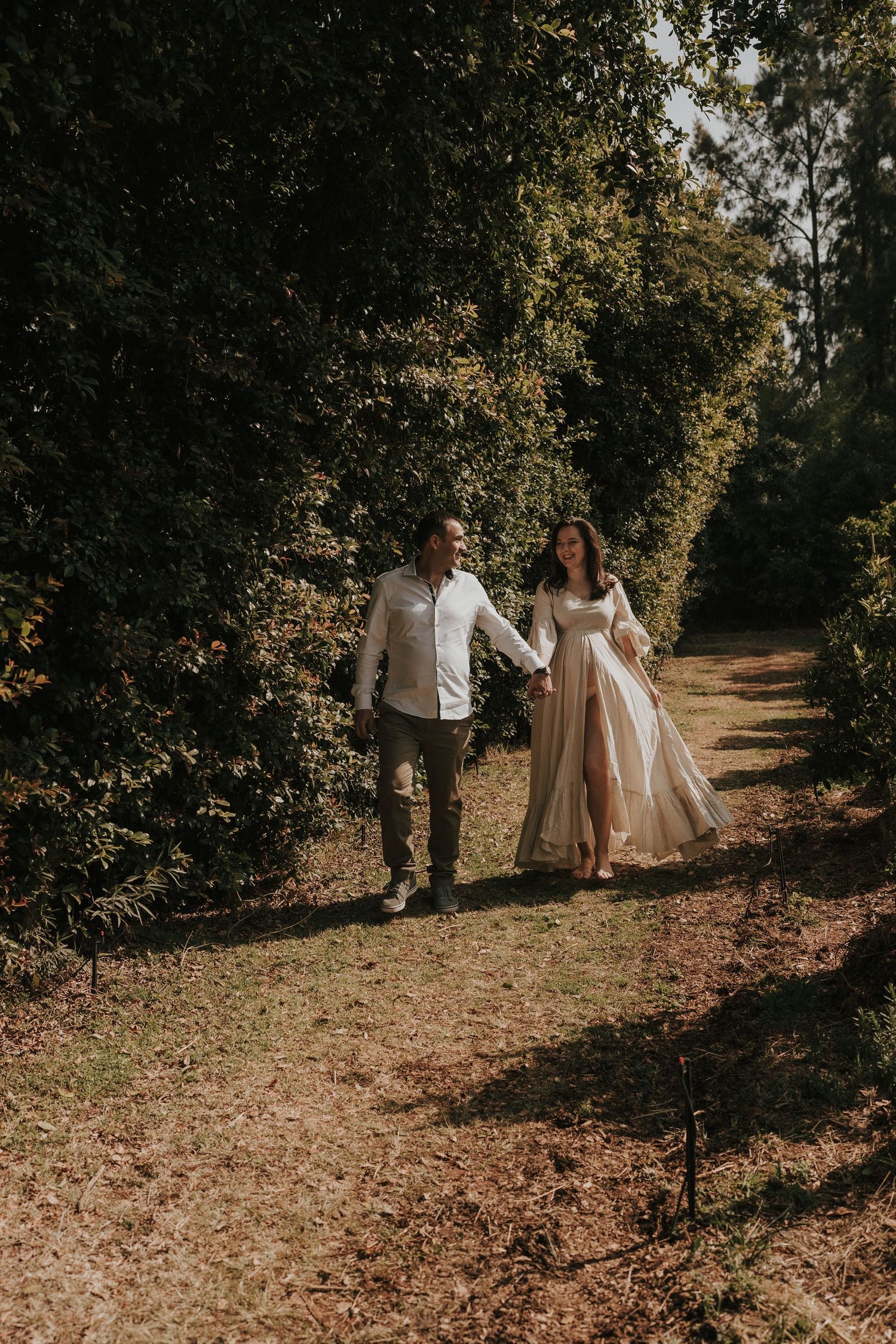 Sun-dappled autumn scene of a couple walking together on a wooded trail in light-colored attire.