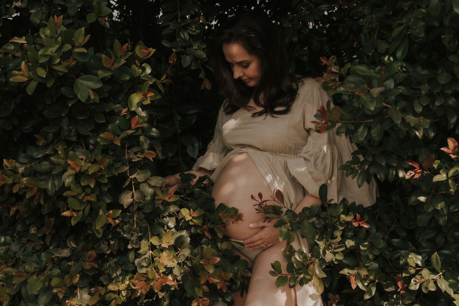 Maternity portrait with golden sunlight filtering through leaves creating a magical garden setting.