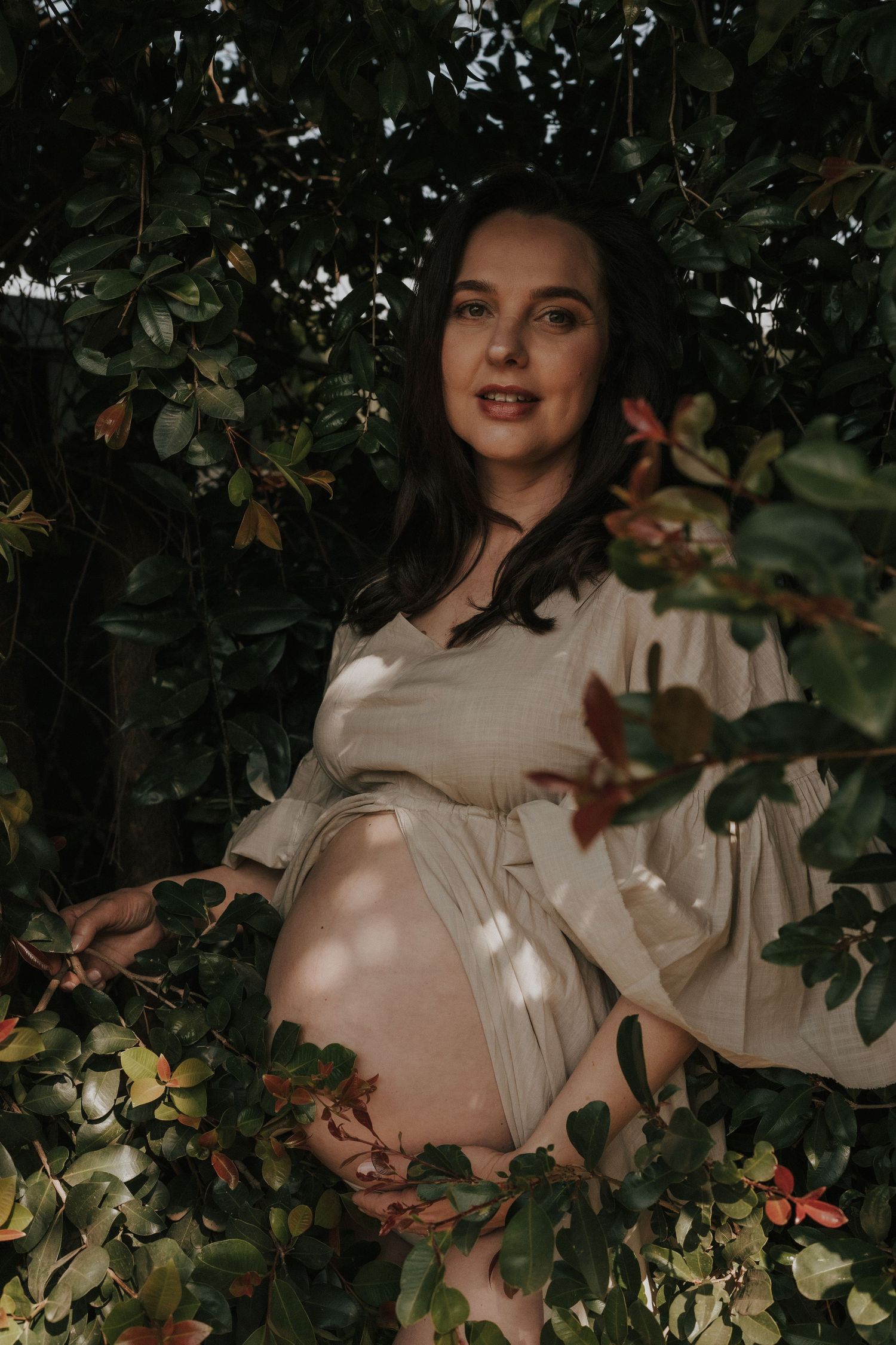 Artistic maternity photo showing a white dress against dark foliage with dappled sunlight.