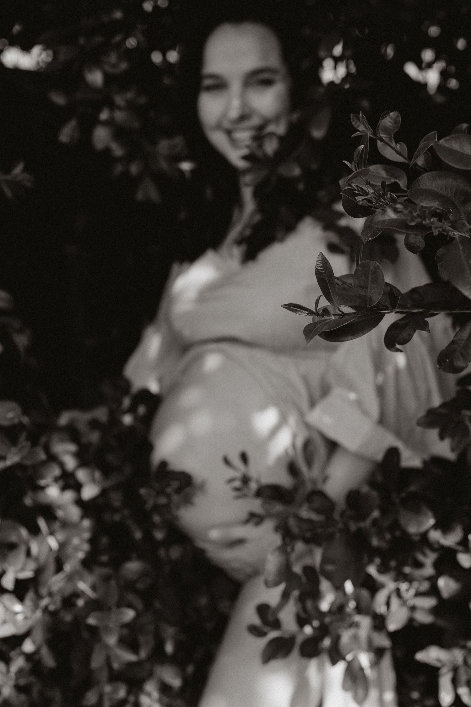 Ethereal maternity silhouette framed by dark leaves and branches.