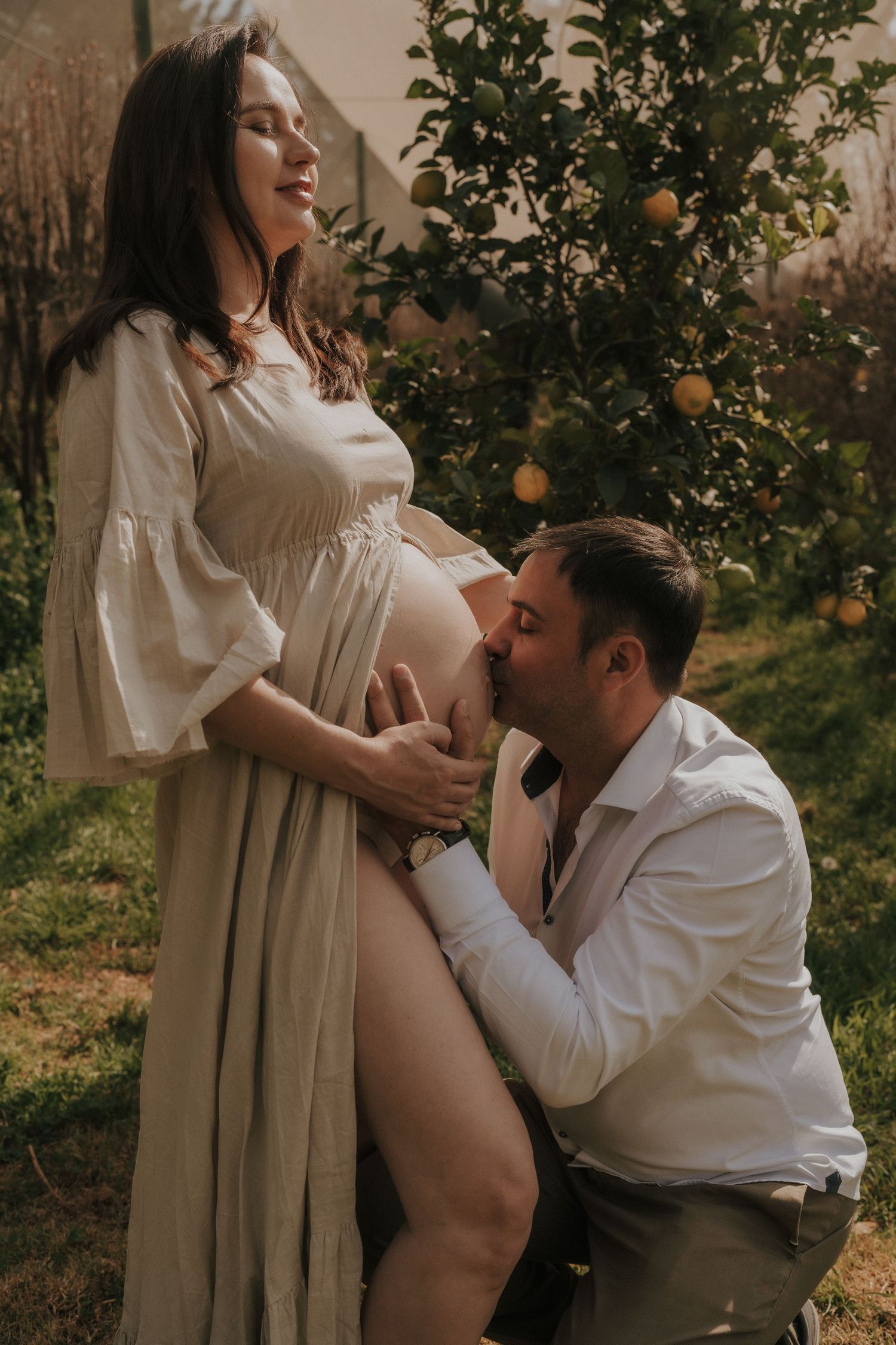 Intimate maternity photoshoot in a citrus grove with flowing white dress and natural lighting.