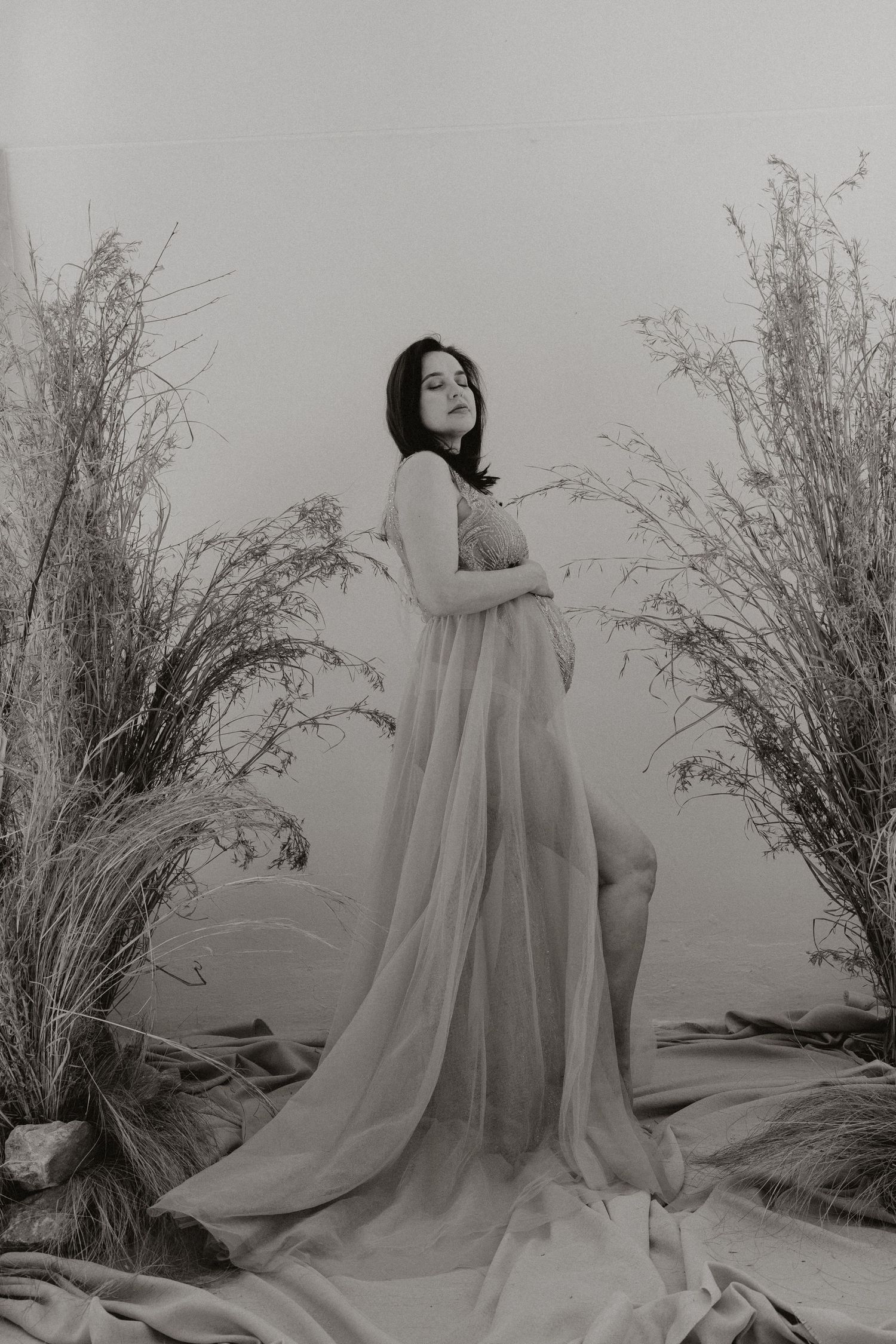 Black and white maternity portrait with dramatic fabric and dried grass elements.