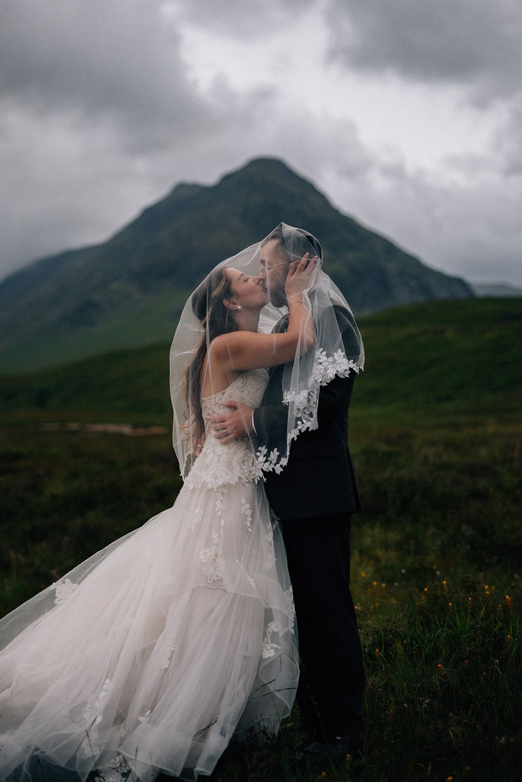 A wedding couple stands by a mountain lake with dramatic peaks in the background.