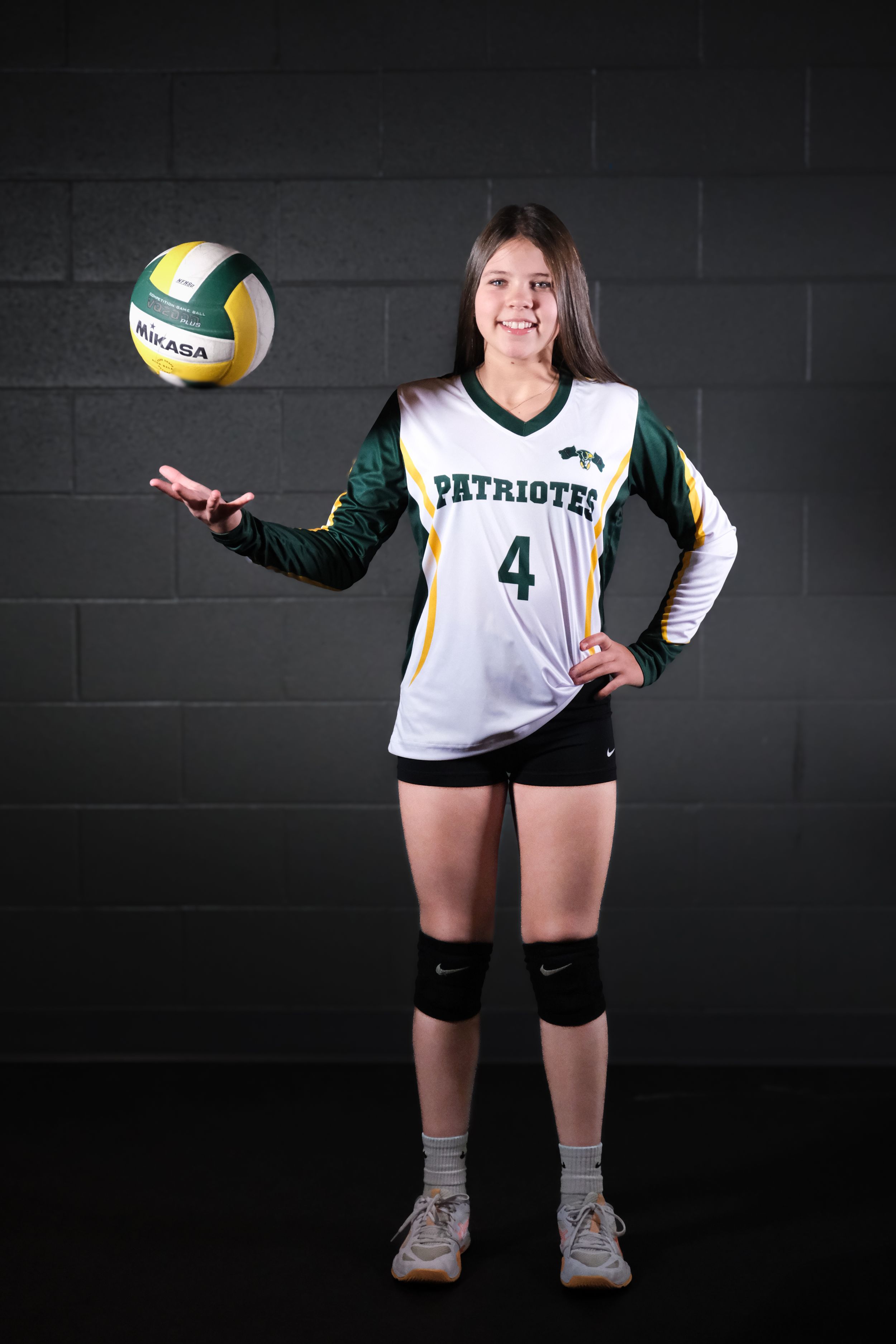 Volleyball player in Patriots uniform number 4 tosses ball against dark background.