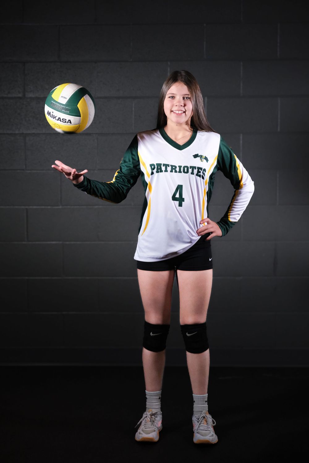 Volleyball player in Patriots uniform number 4 tosses ball against dark background.