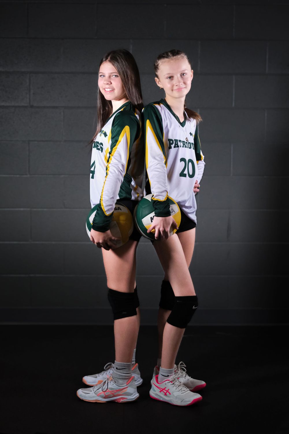 Two volleyball players in Patriots uniforms pose back-to-back against a dark wall.