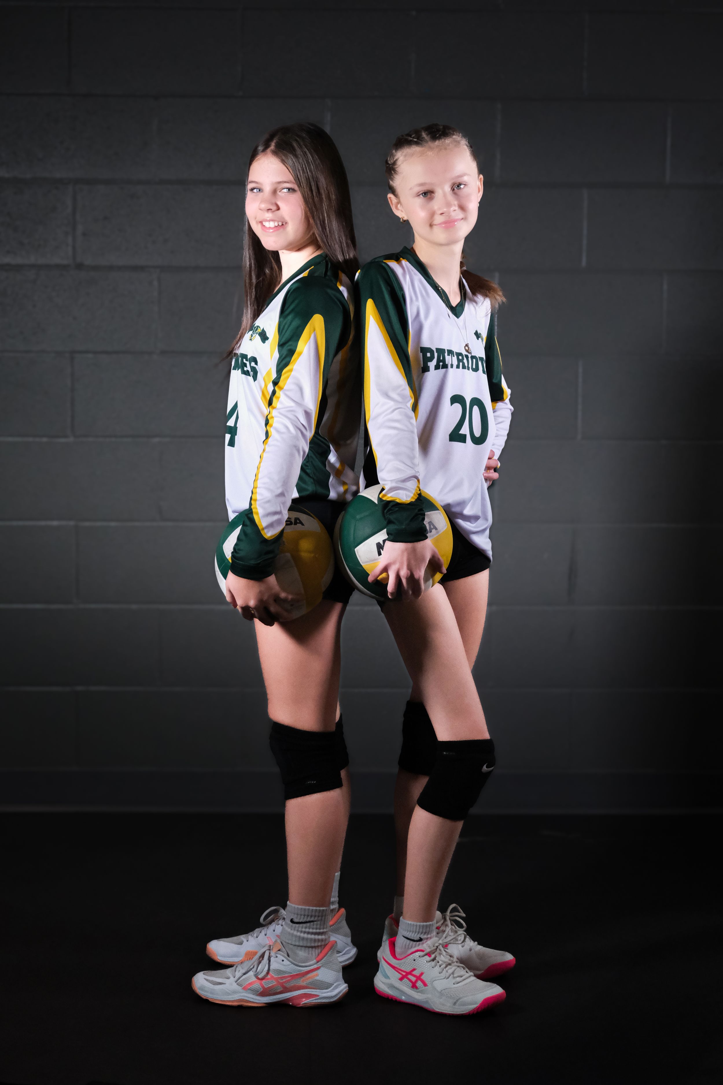 Two volleyball players in Patriots uniforms pose back-to-back against a dark wall.