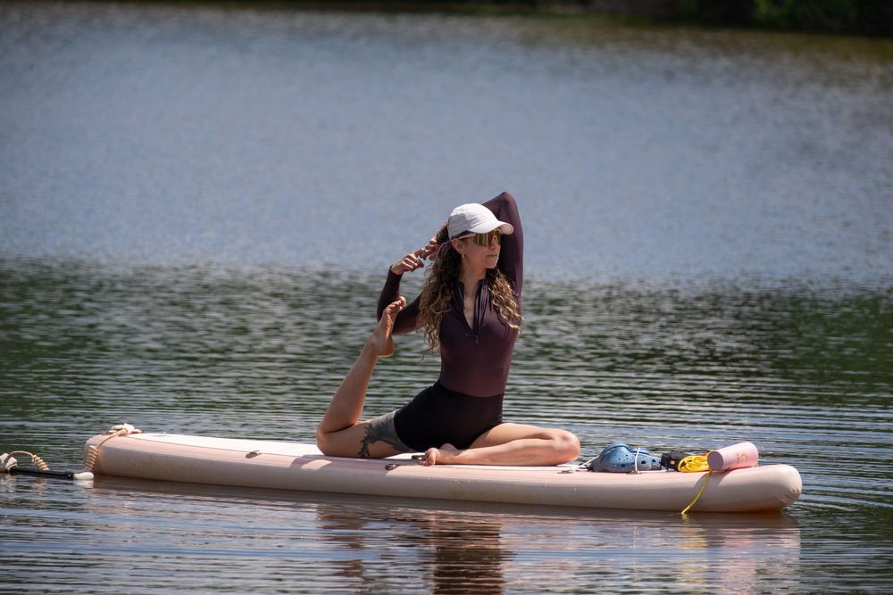 Woman practicing yoga on a paddleboard on a calm lake, wearing a white cap and dark athletic wear.