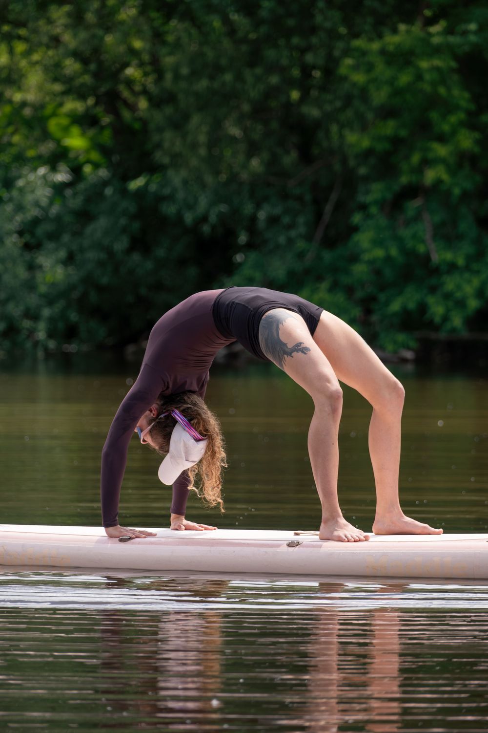 Woman performing a backbend yoga pose on a paddleboard on a calm river surrounded by lush green trees.