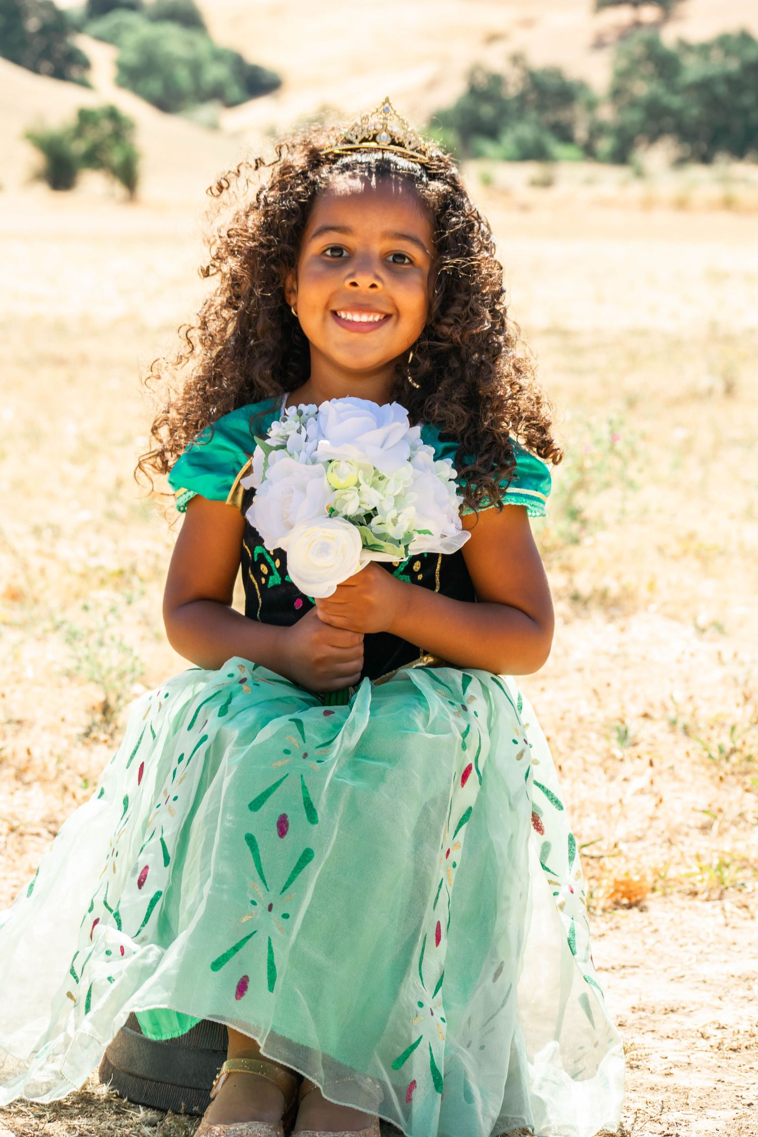 A child in a mint green dress with floral details holds a bouquet of white flowers in a sunny outdoor setting.
