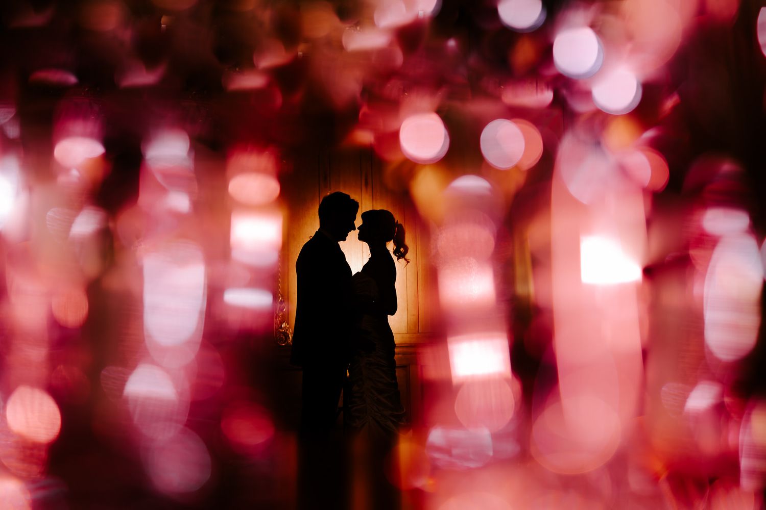 Silhouetted figures dance against a vibrant red and pink bokeh light background creating a romantic atmosphere.