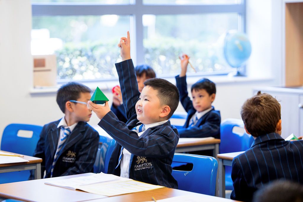 Students raise their hands enthusiastically to participate during class in a bright modern classroom setting.