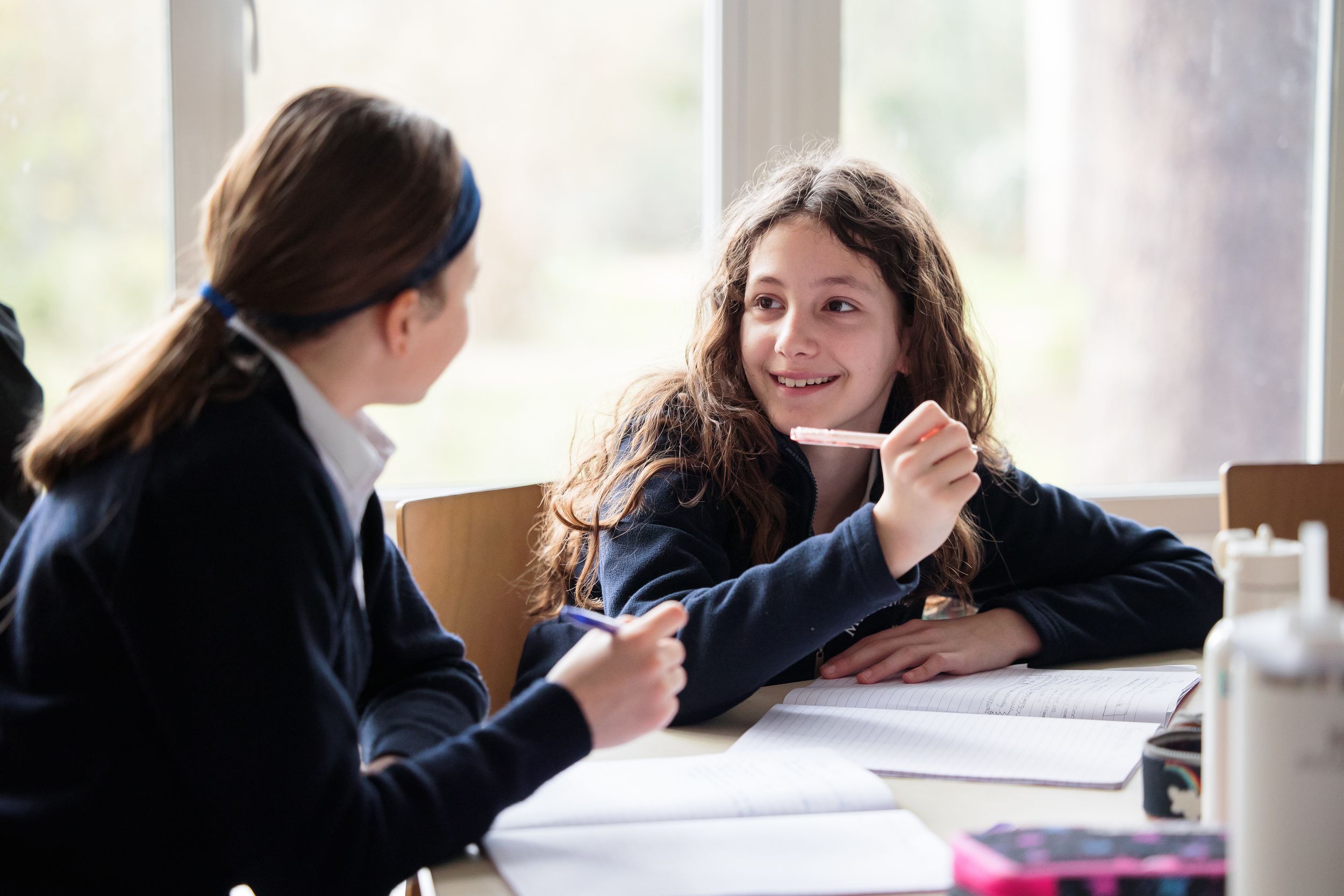Two students engage in conversation while studying at a bright window-lit table.