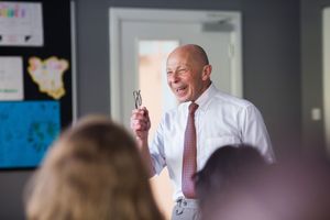 A speaker gestures enthusiastically while presenting to an audience in a classroom setting.