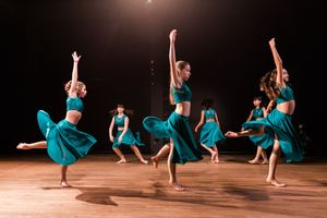 Dancers in turquoise dresses perform a synchronized modern dance routine on a wooden stage.