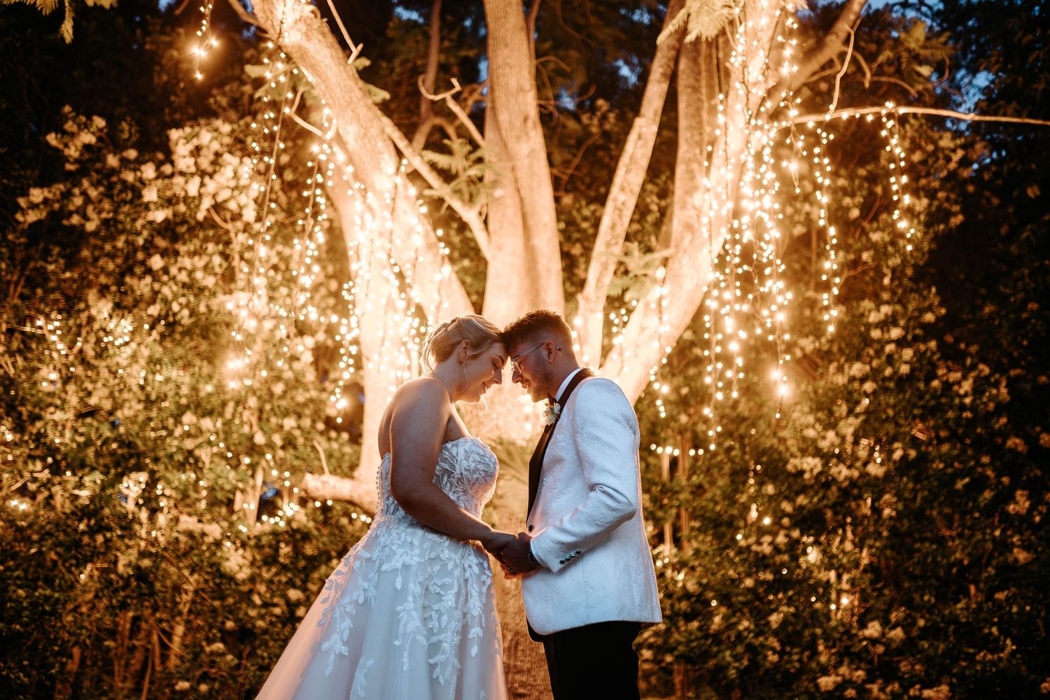 Romantic couple shares an intimate moment under twinkling string lights in a garden setting at dusk.