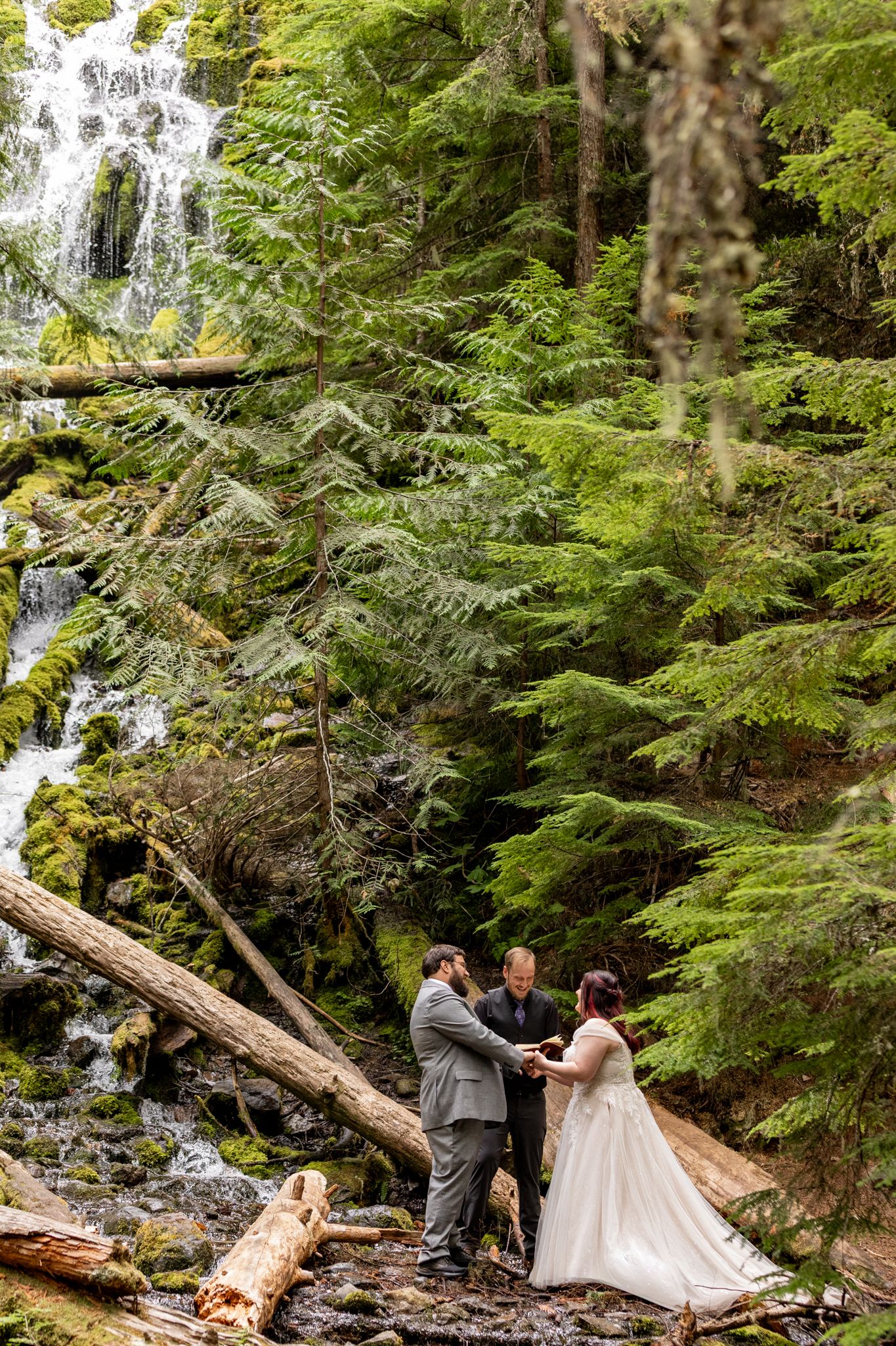 Bride and groom exchanging vows with officiant beside a stunning waterfall in a lush green forest. PNW elopement Photographer.