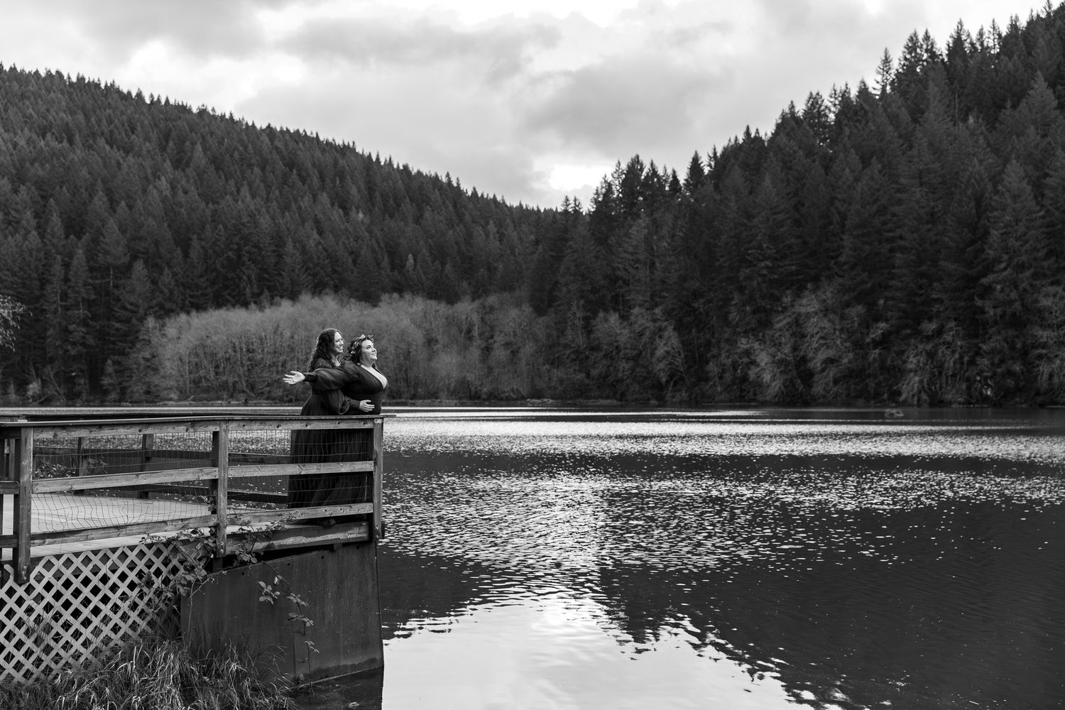 Couple standing on a wooden dock overlooking a calm lake surrounded by dense evergreen forest in black and white. PNW elopement Photographer