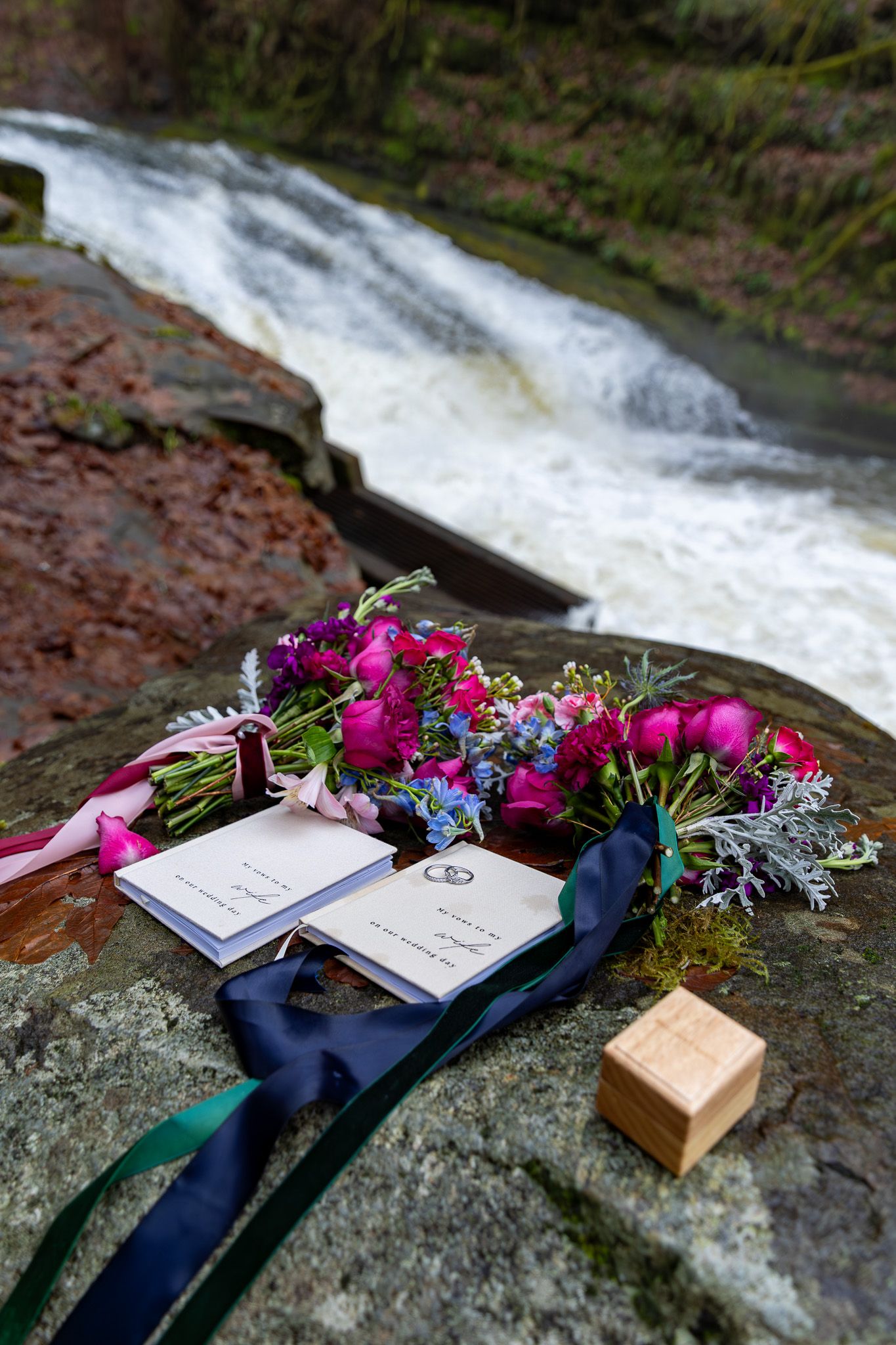 Wedding bouquets, vow books, and rings displayed on mossy rocks near waterfall. PNW elopement Photographer.