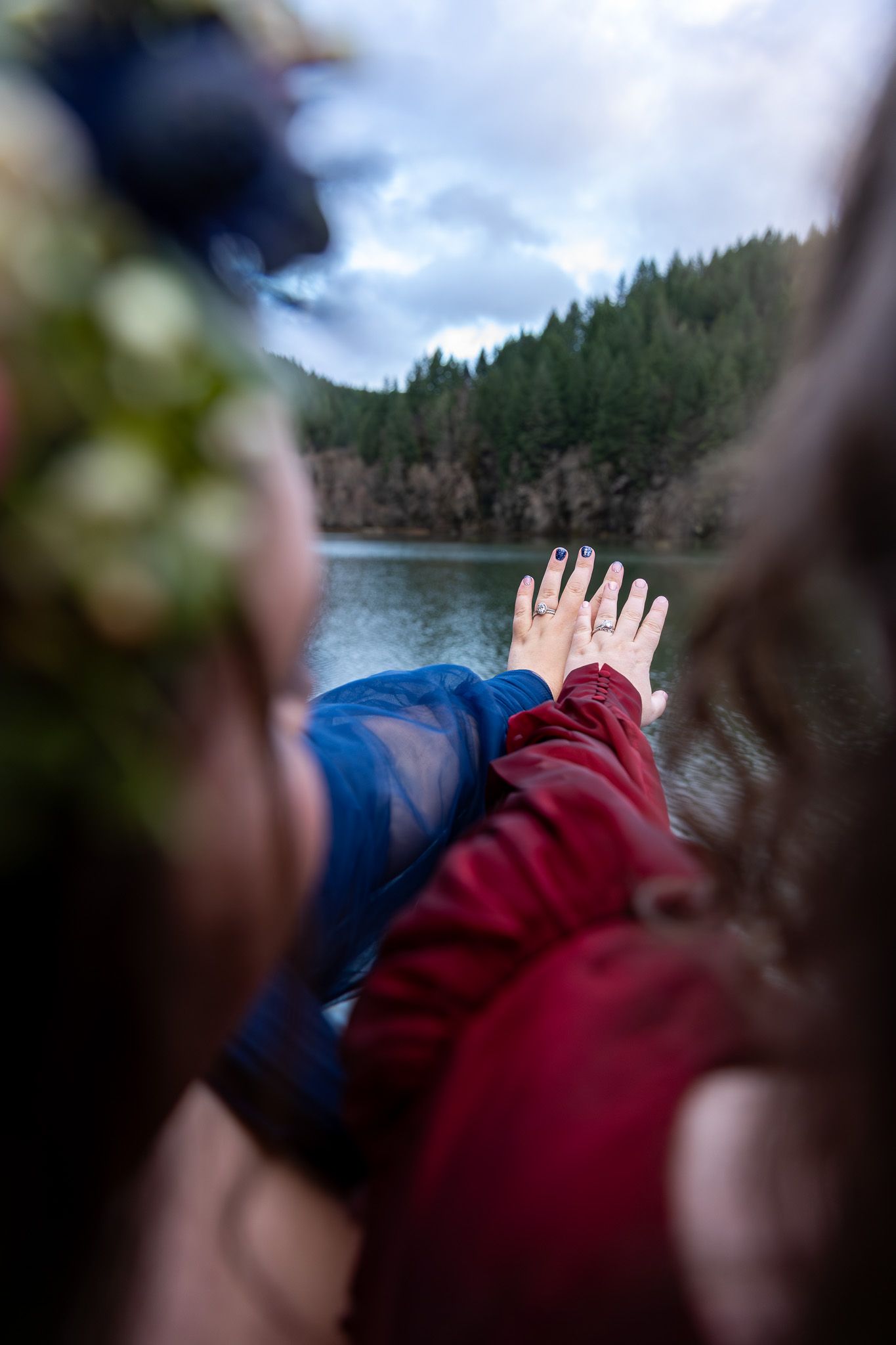 Close-up of two brides holding hands showing wedding rings overlooking a serene lake. PNW elopement Photographer.