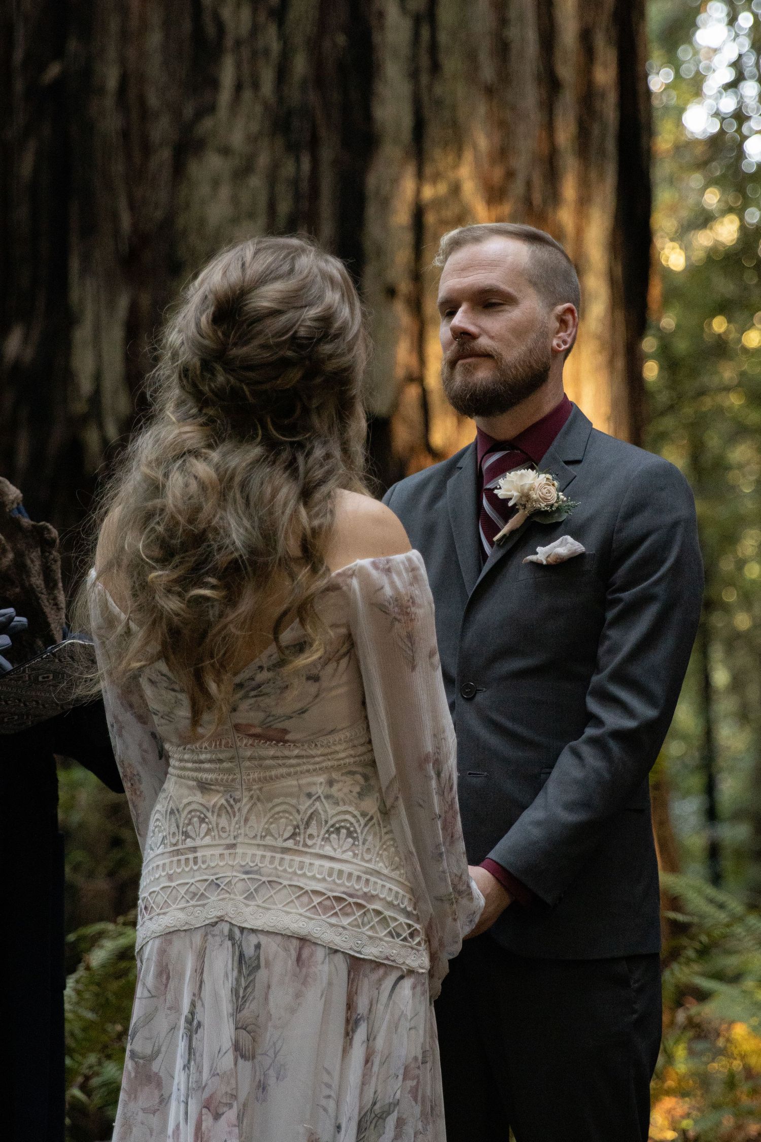 Groom in gray suit holding bride's hands during forest wedding ceremony among giant redwood trees.