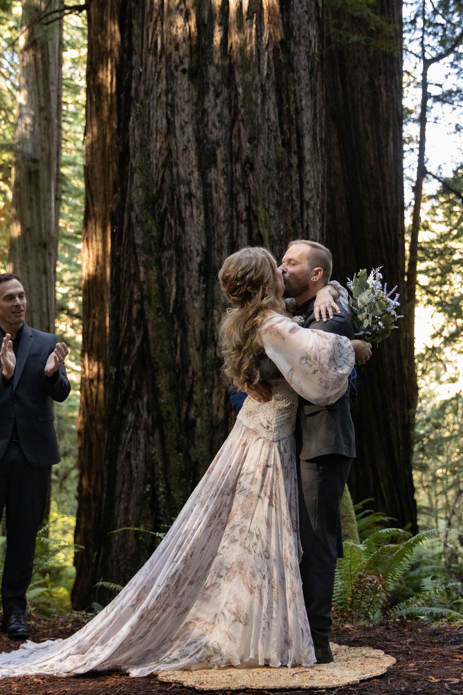 Newlywed couple shares first kiss embrace surrounded by giant redwood trees in forest wedding.