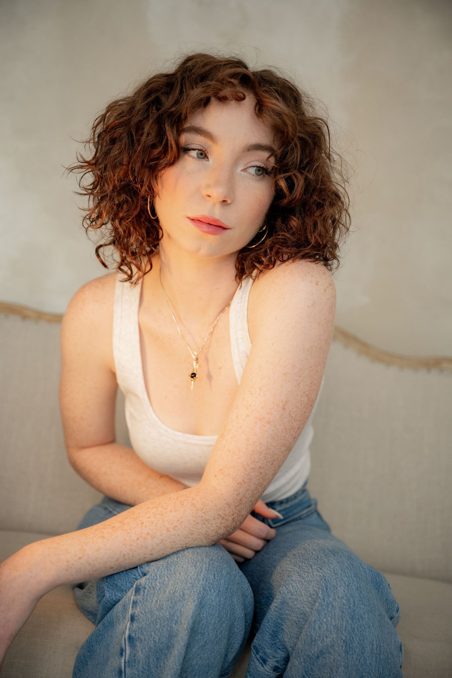 A person with curly hair and red lipstick wearing a white tank top and jeans poses against a neutral background.