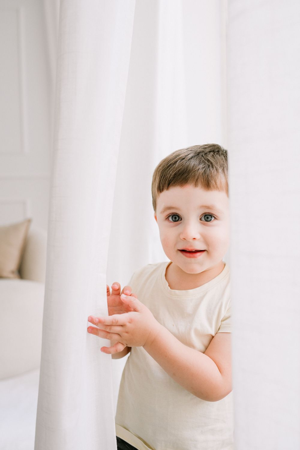 Joyful toddler clapping hands while playing in front of white curtains.