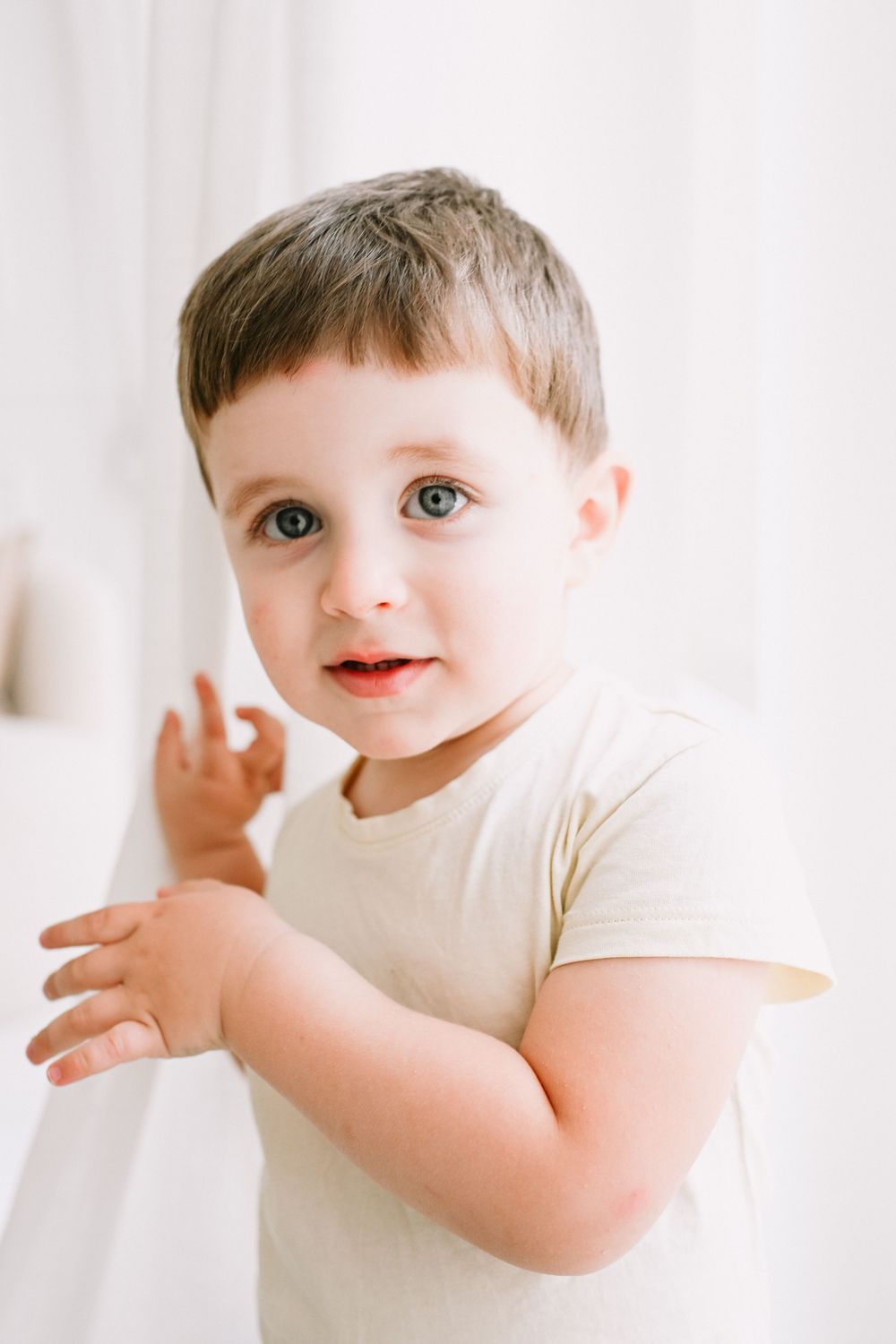 Playful toddler making expressive gestures against white curtain backdrop.