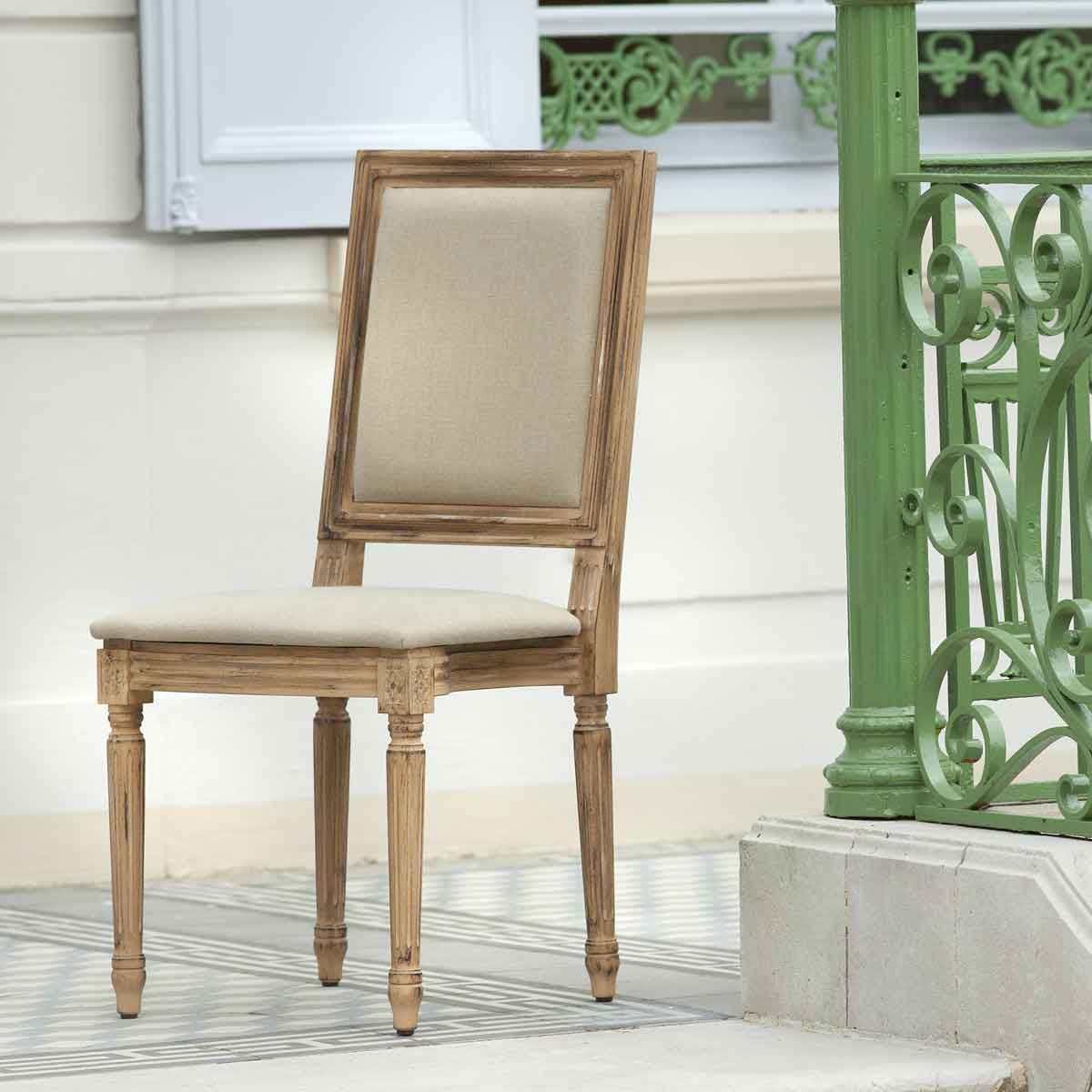 Élégante chaise de campagne française avec rembourrage beige et cadre en bois de chêne patiné sur un porche près de la balustrade verte.