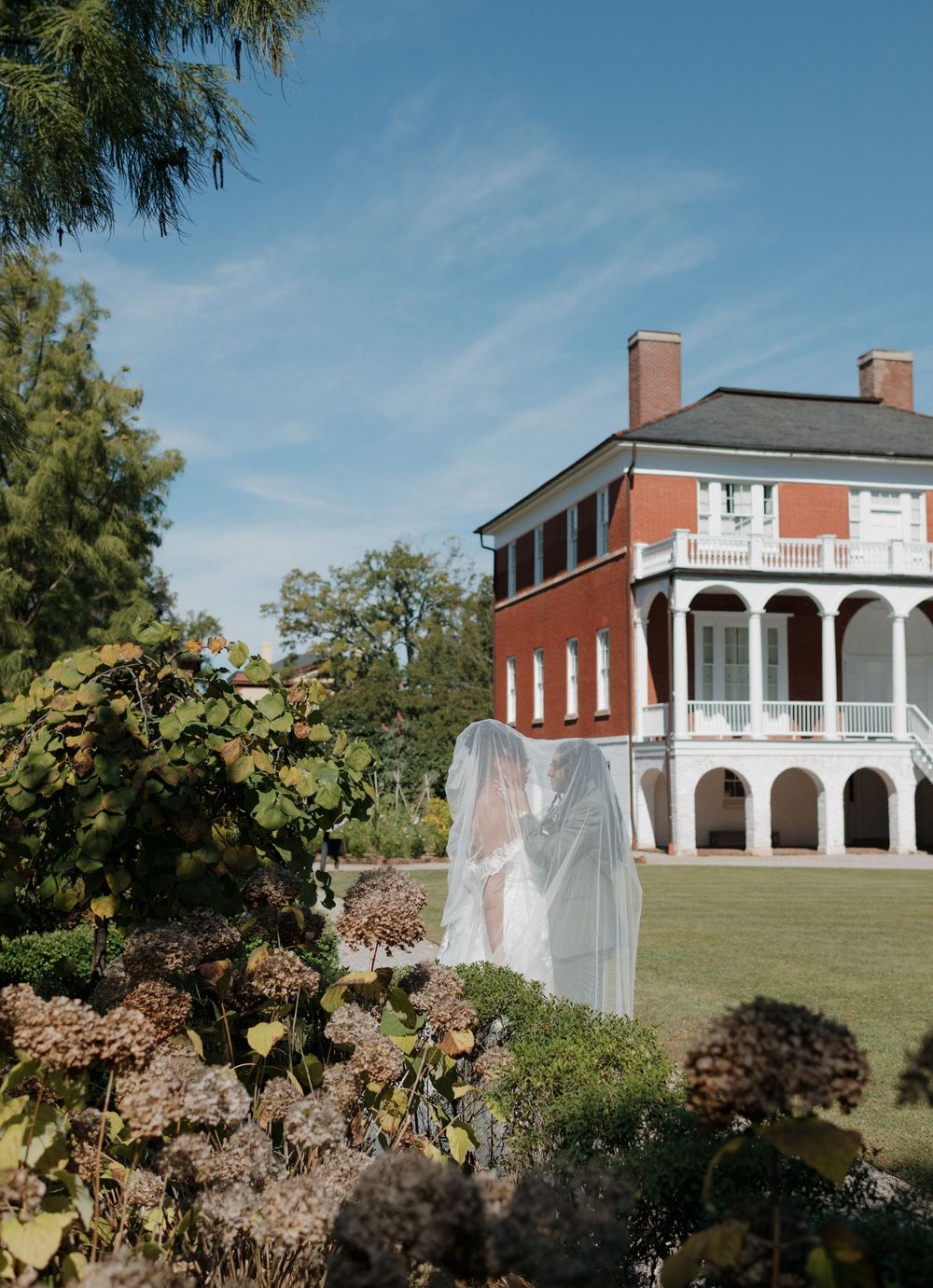 Historic brick mansion with white columns and wraparound porch sits atop a grassy hill surrounded by autumn foliage.