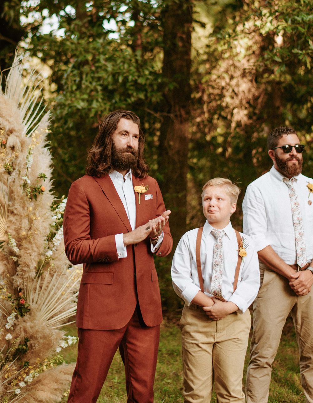 Wedding party members in rust-colored suits and beige attire pose together in an outdoor garden setting.