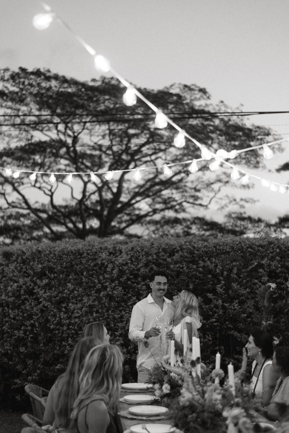 Black and white outdoor garden party scene with string lights and people gathered around a table at dusk.