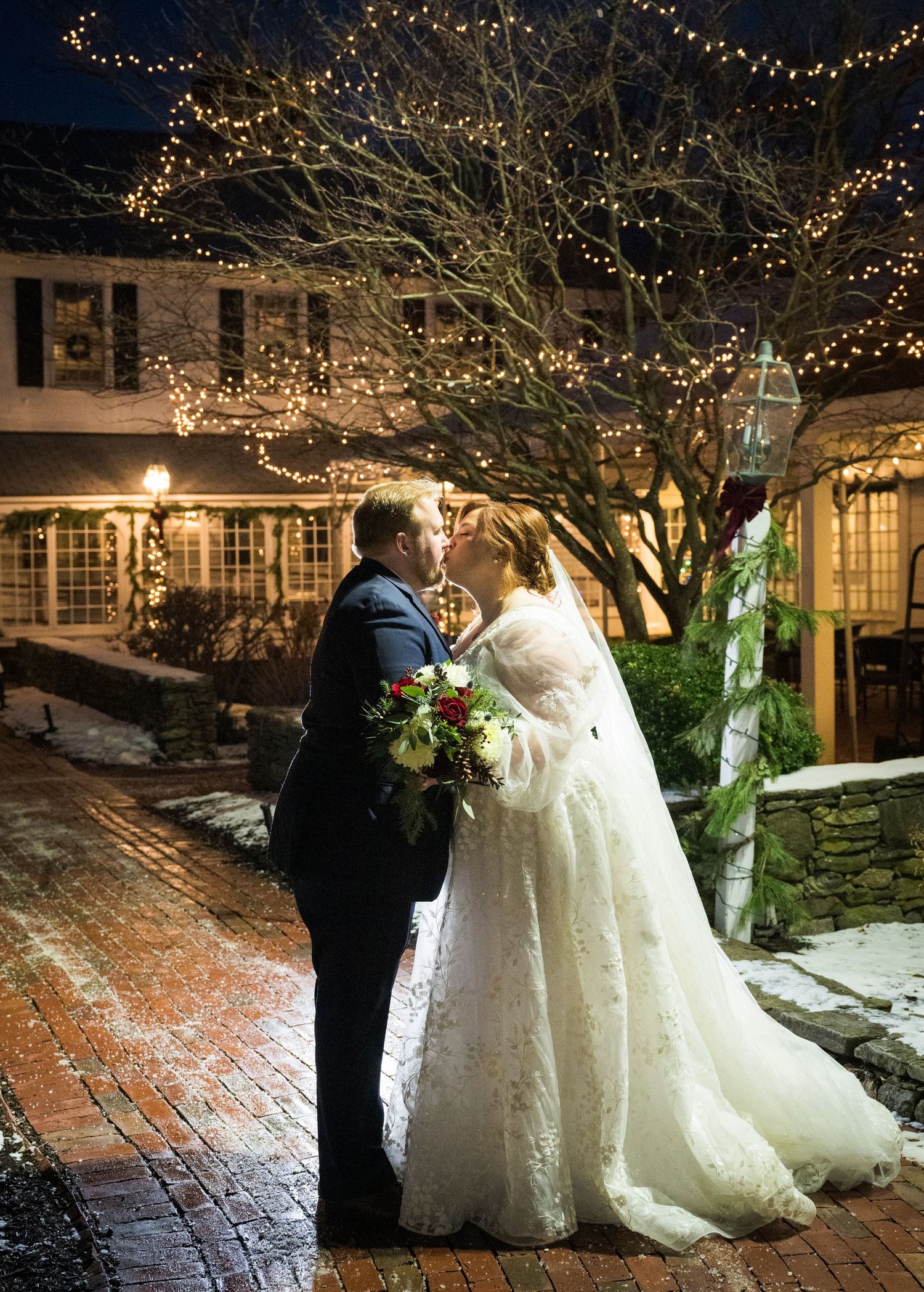 A romantic winter wedding scene with a couple embracing under a tree decorated with twinkling lights on a snowy evening.