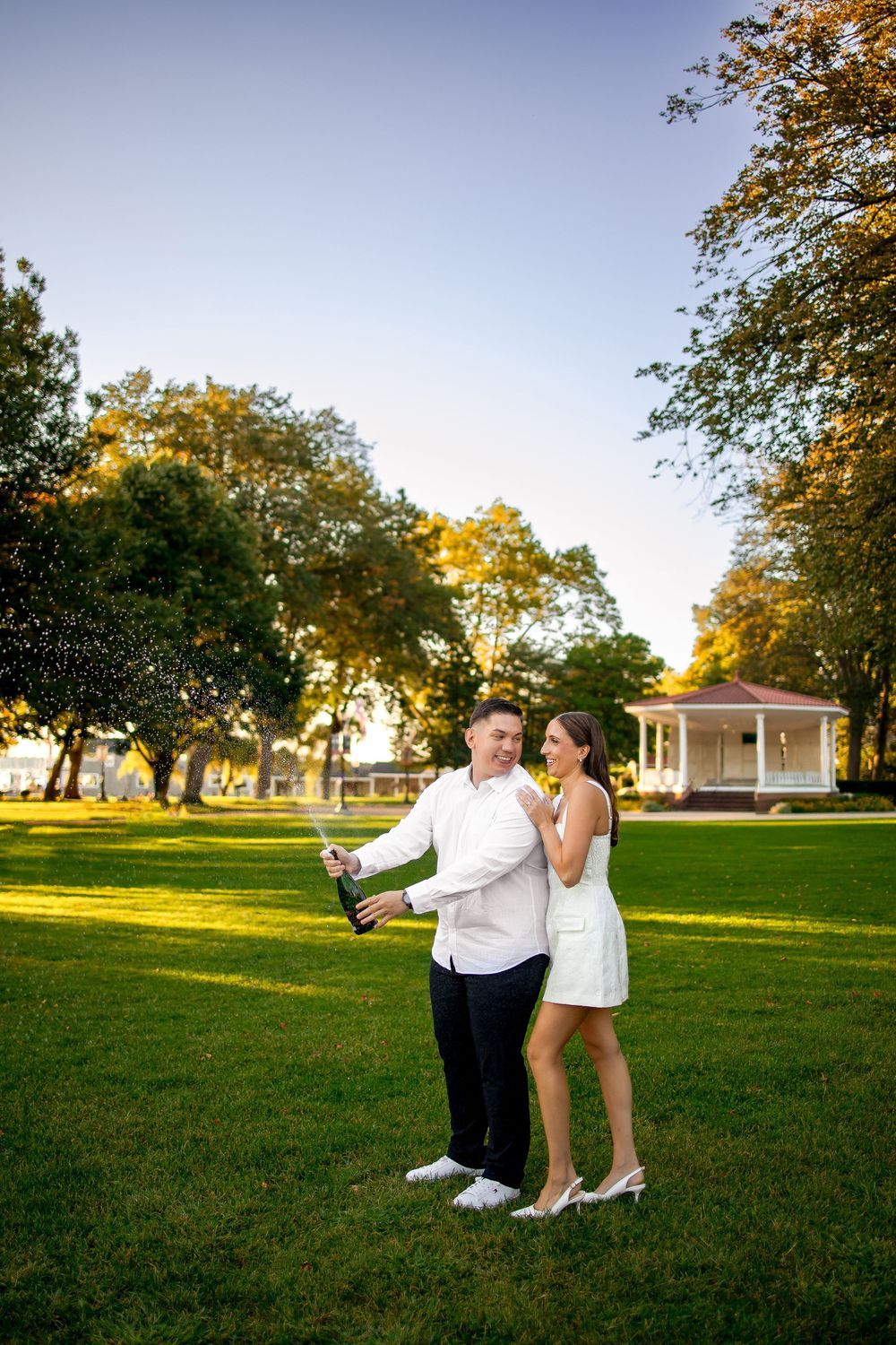 A couple poses together on a grassy lawn at sunset with trees and buildings in the background.