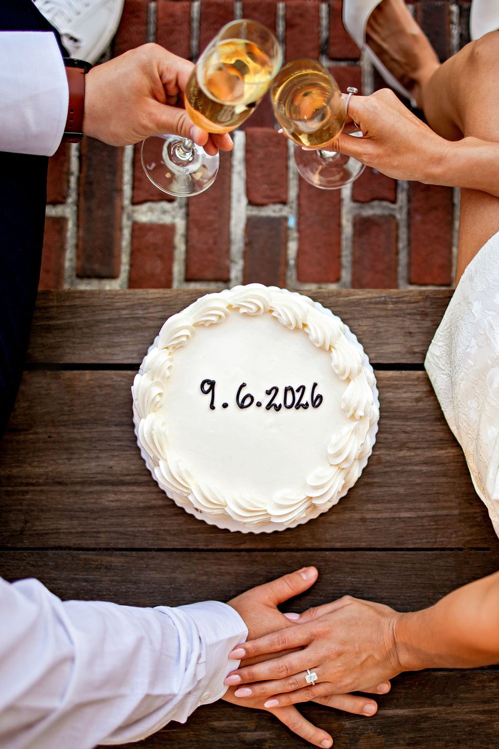 Hands reach across a wooden table with wine glasses and a white plate displaying a wedding date of 9.6.2026.