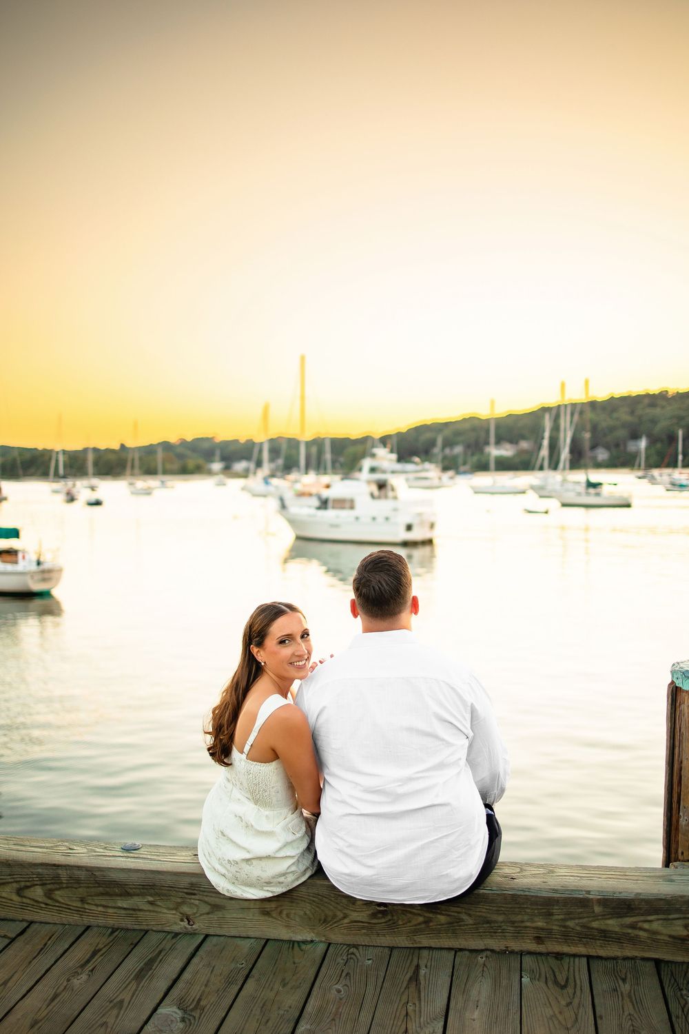 A couple sits together on a wooden dock overlooking a marina filled with sailboats during golden hour.