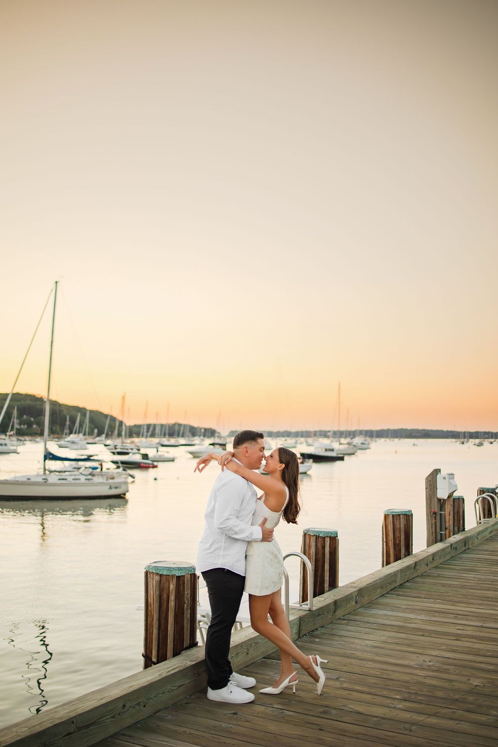 Romantic couple shares tender moments on a wooden pier at sunset with sailboats in the marina background.