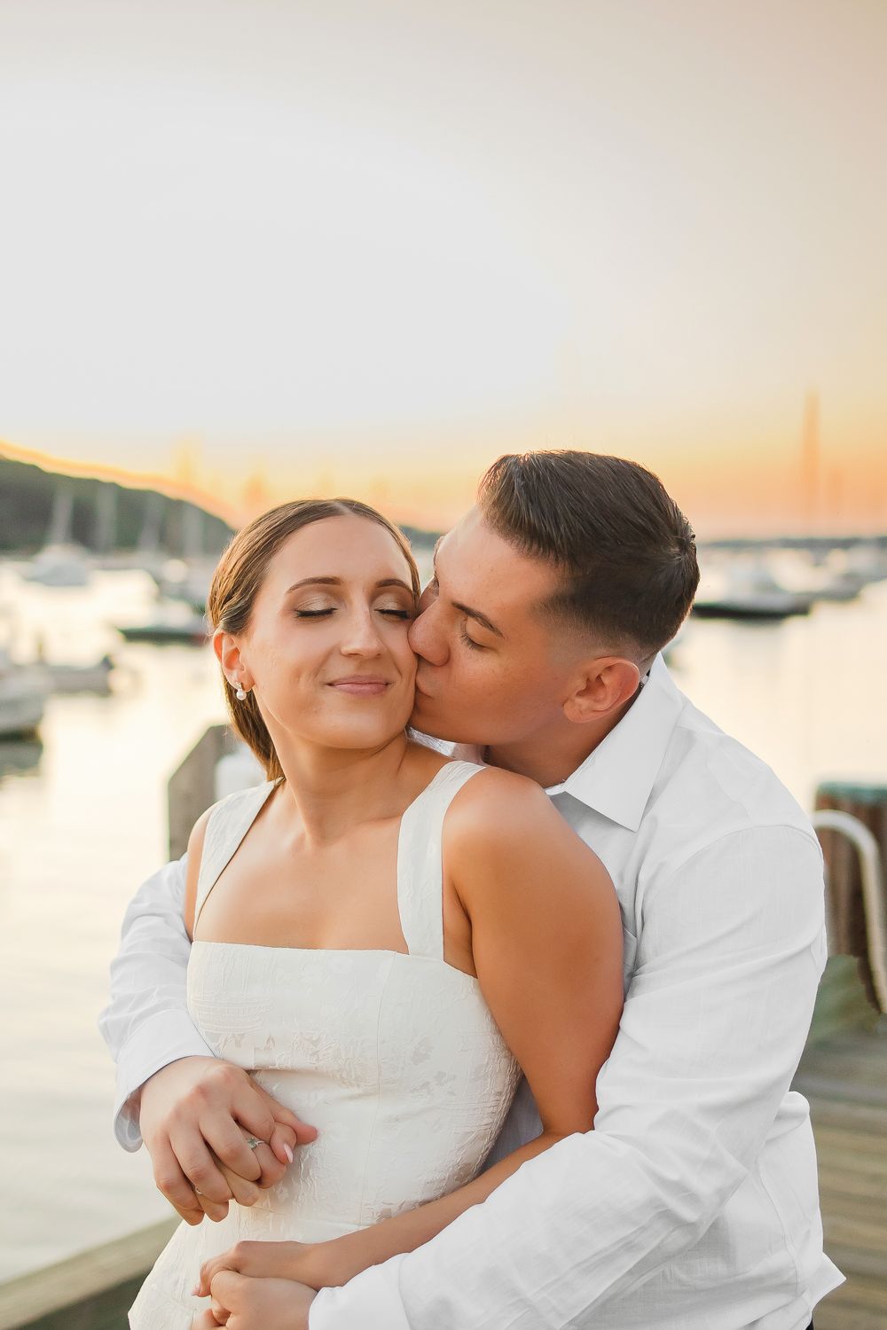 A couple shares an intimate moment on a dock at sunset with boats in the marina background.