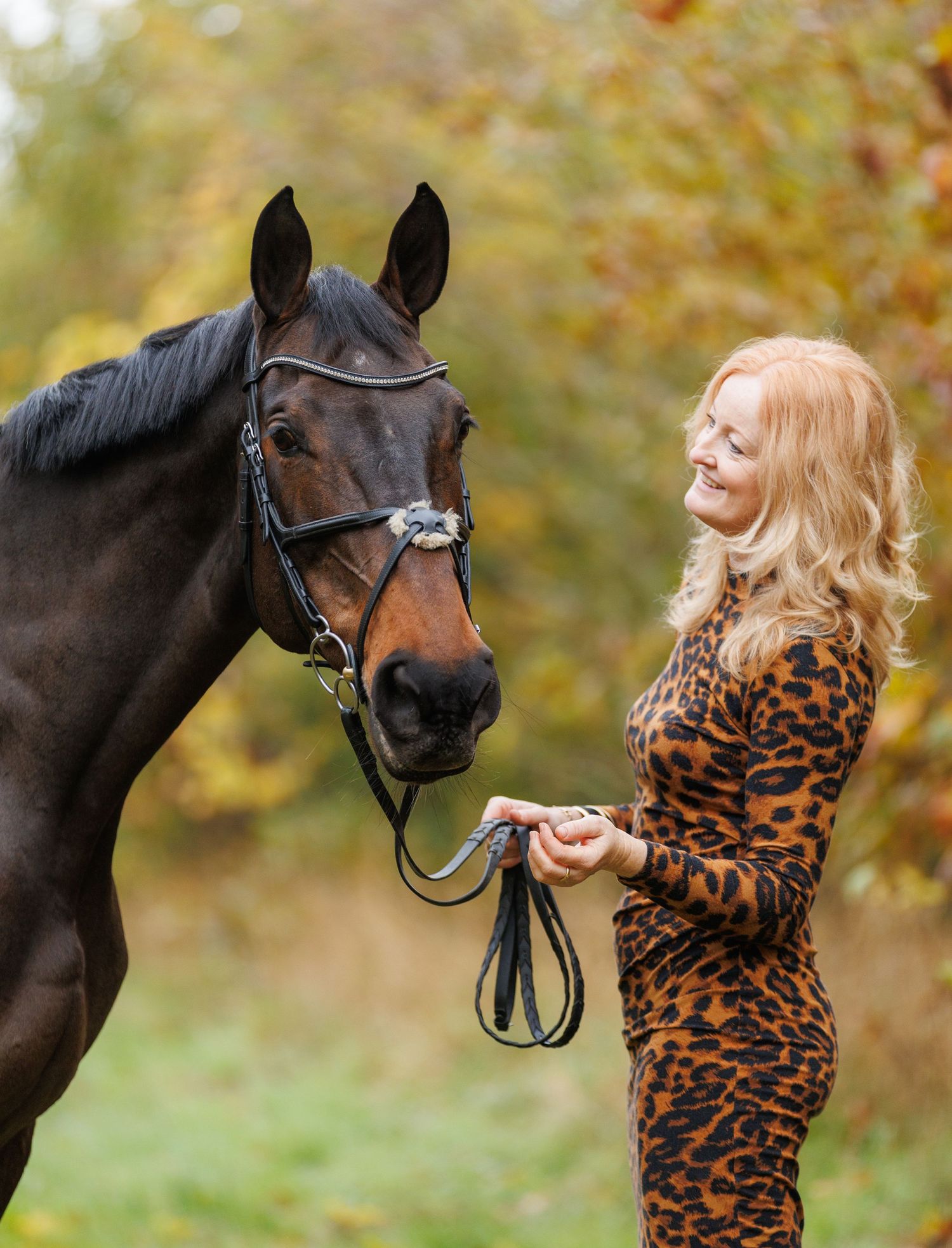 A brown horse and rider in a white sweater pose together against autumn foliage in soft natural lighting.