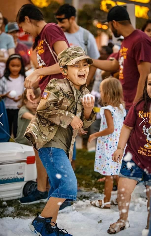 A young person in camouflage and overalls dances joyfully at an outdoor summer gathering with a crowd in the background.